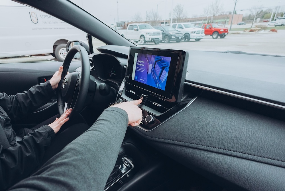 Hazard-button-in-vehicle-DriveWise A person sits in the driver's seat of a car, holding the steering wheel with one hand and using the touchscreen display on the center console with the other hand. Several parked vehicles are visible outside.