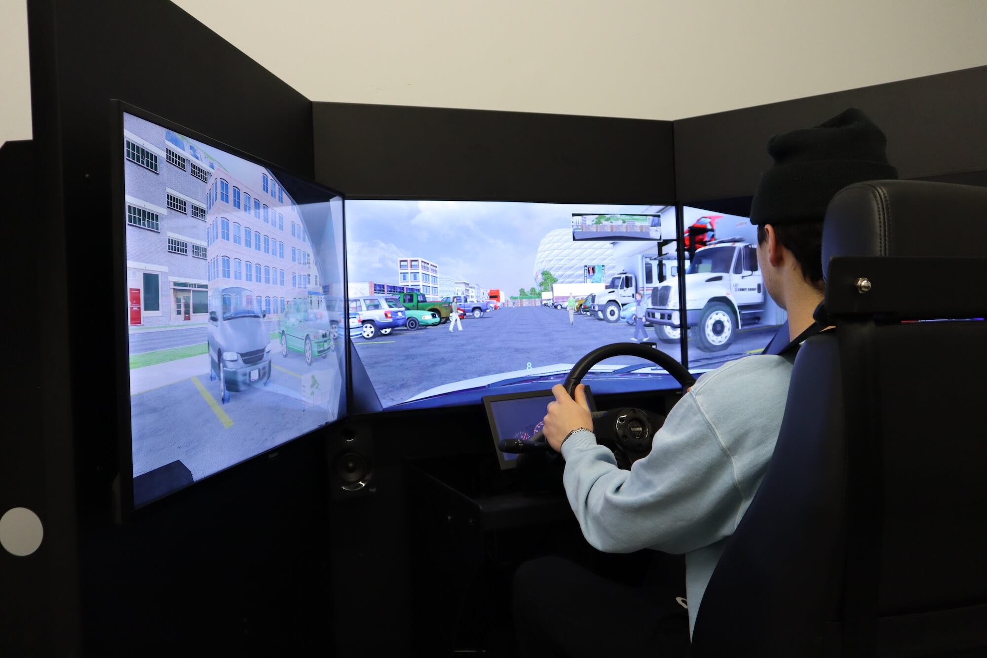 Driving-On-Simulator-DriveWise-Newmarket A person sits in a driving simulator with three large screens showing a virtual urban street scene with vehicles, pedestrians, and buildings. The person holds a steering wheel and looks at the central screen.