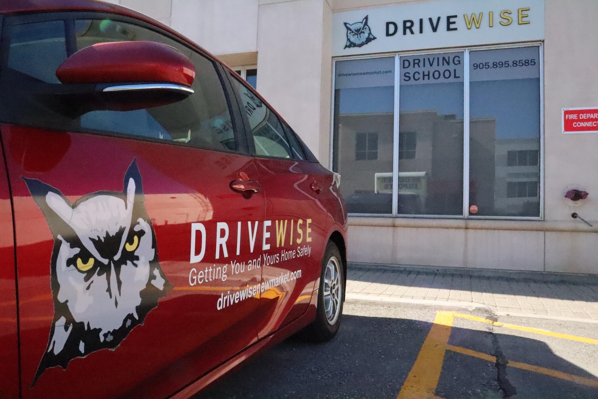 Outsid-View-DriveWise-Newmarket A red car with a DriveWise logo featuring an owl is parked in front of a building with a DriveWise Driving School sign, a phone number, and a red fire department connection notice on the wall.