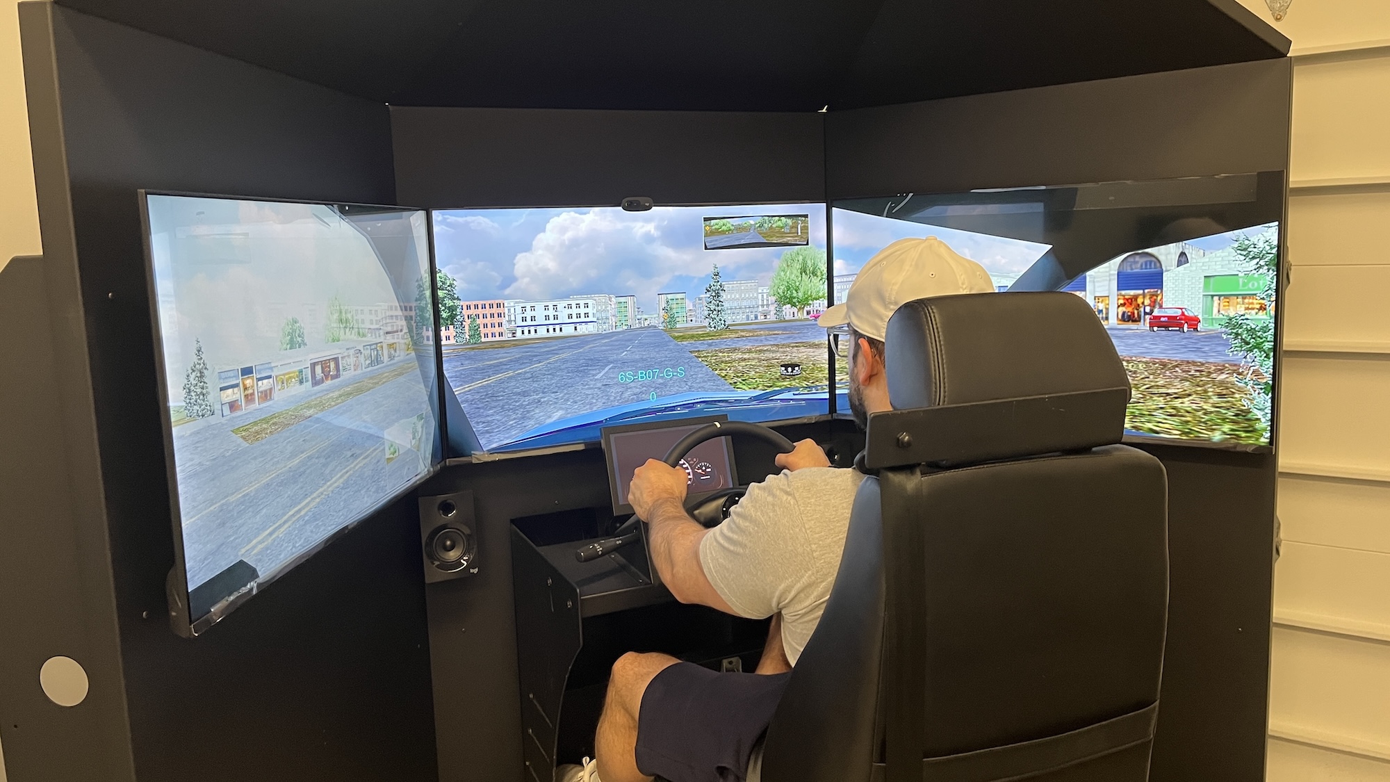 A person wearing a white cap and gray shirt is seated in a driving simulator with three large screens displaying a city street scene and holding the steering wheel as if driving.