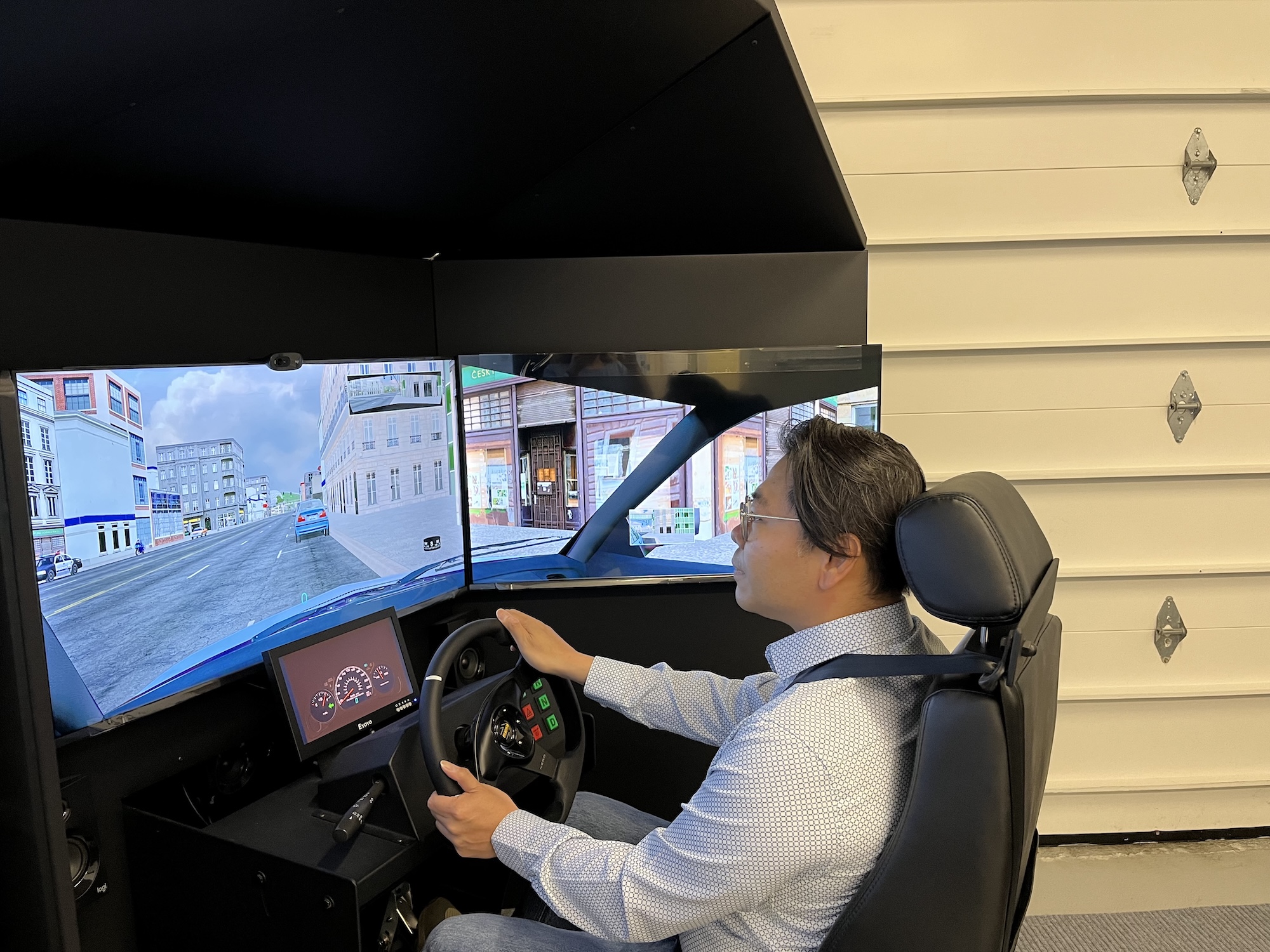 DriveWise-Newmarket-simulator-driving-lessons A person sits in a driving simulator, holding the steering wheel and looking at three screens displaying a virtual city street scene. The simulator equipment includes a dashboard and various controls.