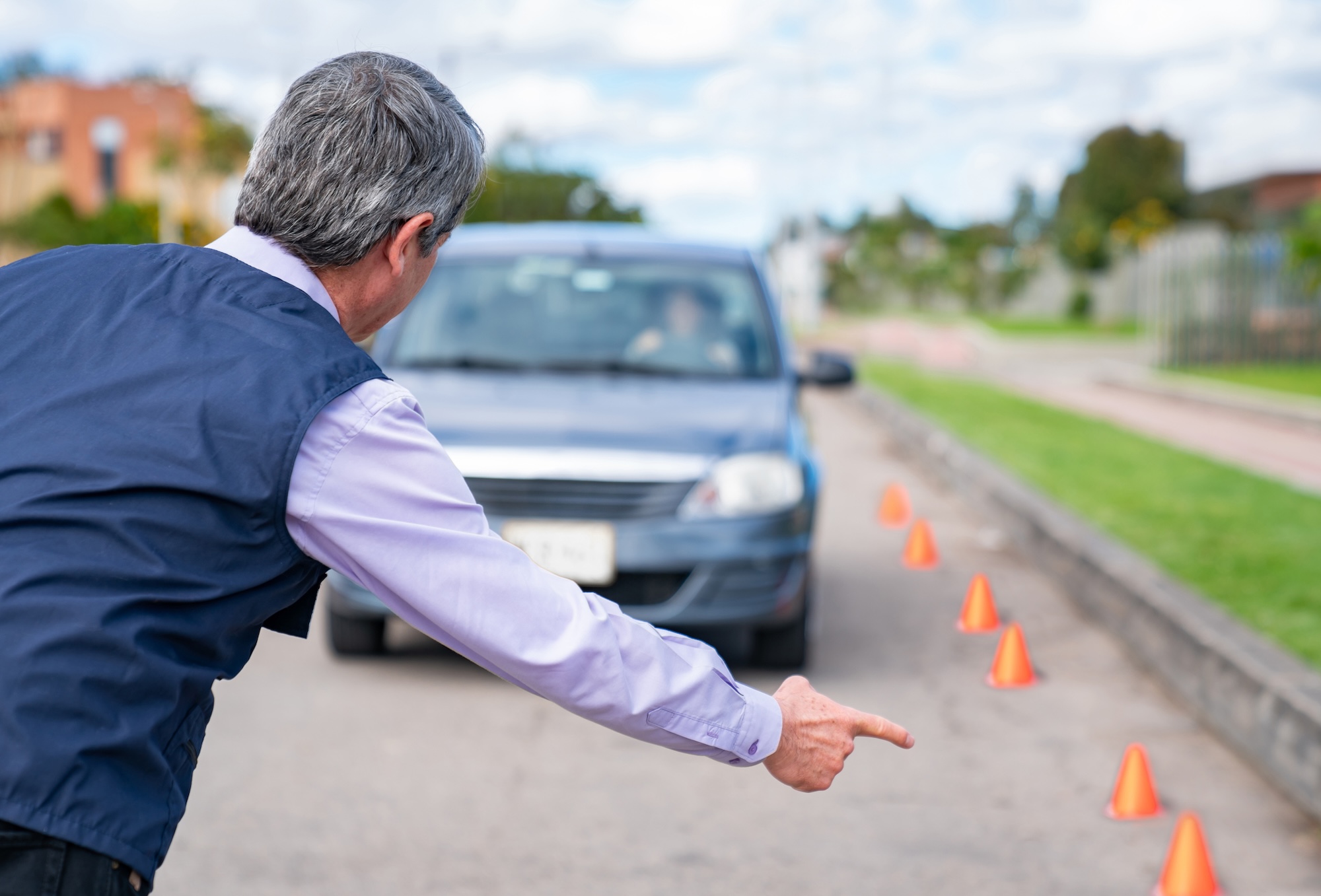Driving instructor giving instructions to a woman taking driving lessons An instructor wearing a vest points toward a line of orange traffic cones on a paved road, guiding a person driving a blue car through a driving test or training exercise outdoors.