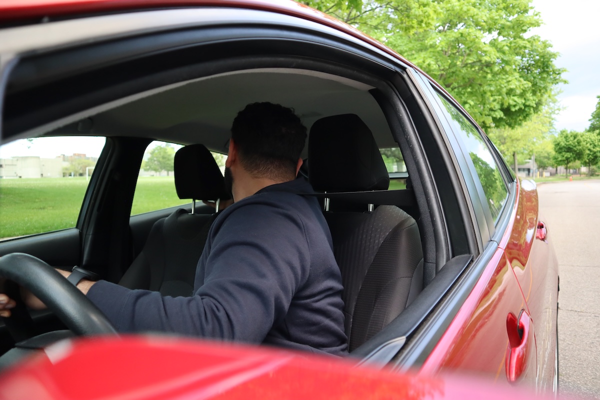 Driver-reversing-DriveWise-Newmarket A person in a dark hoodie sits in the driver's seat of a red car, looking backward over their shoulder, likely preparing to reverse. The car is parked on a street next to a grassy area with trees.