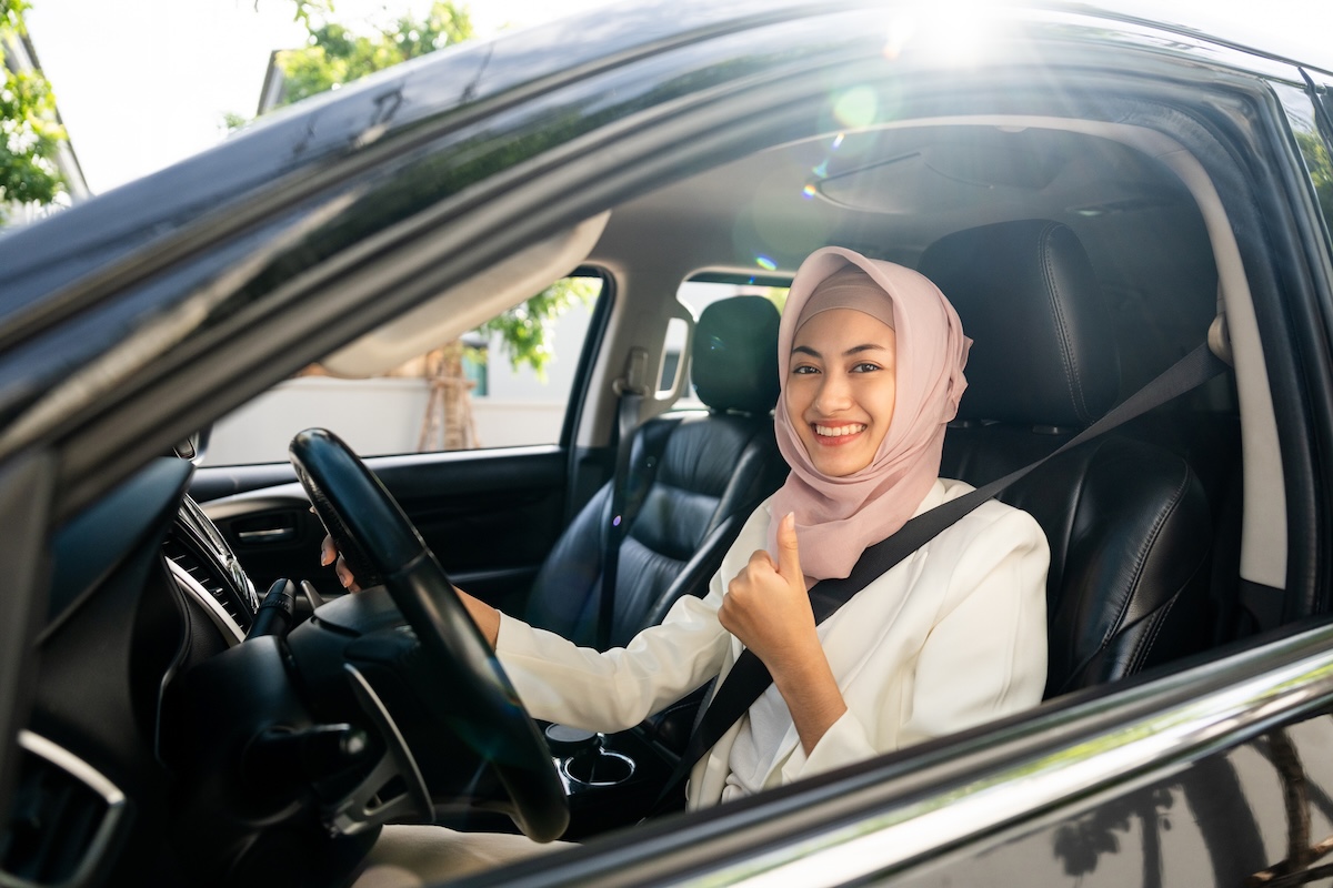 Asian muslim business women getting new car. She very happy and excited. Smiling female islamic driving vehicle on the road on a bright day with sun light. Business woman buying driving new car A woman wearing a light pink hijab and white outfit sits in the driver's seat of a car, smiling and giving a thumbs-up while looking out the open window. Sunlight streams into the vehicle.