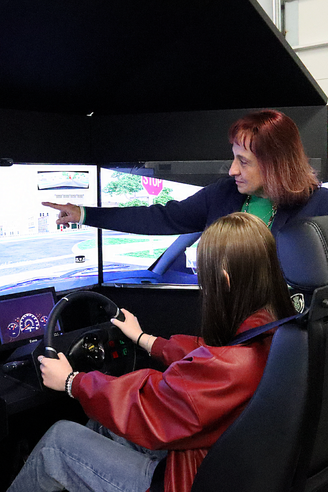 Fida-SimulatorPointing An instructor stands beside a seated person holding a steering wheel in a driving simulator. The instructor points at the simulation screen displaying a street scene and traffic signs.