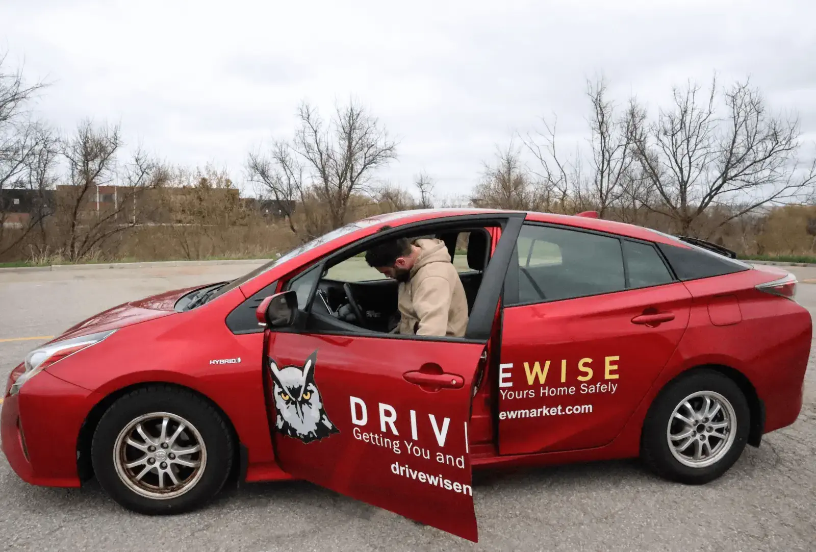 Getting-In-Car-DriveWise-Newmarket A person in a beige hoodie sits in the driver's seat of a red car with "DRIVEWISE" and an owl logo on the side, parked on a paved surface near leafless trees under a cloudy sky.