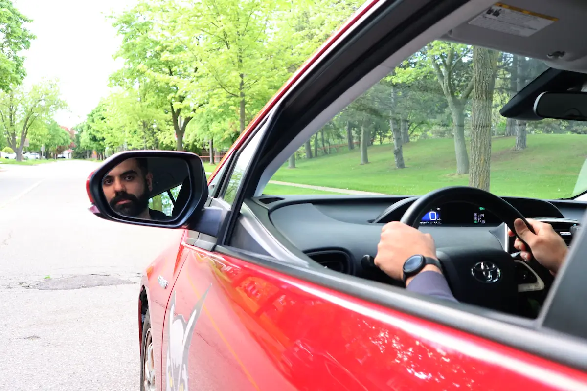 New-Canadian-Driver-DriveWise-Newmarket A person sits inside a red car, holding the steering wheel, with their reflection visible in the driver’s side mirror. The car is parked on a tree-lined street with green grass and foliage outside.