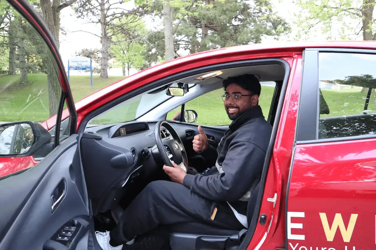 New-Canadian-Driver-with-thumbs-up-DriveWise-Newmarket A man sits in the driver’s seat of a red car with the door open, smiling and giving a thumbs-up. Trees and grass are visible outside, and part of the car’s lettering is shown on the door.