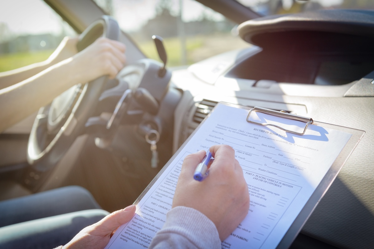 Examiner filling in driver's license road test form A person holding a clipboard with a form while another person sits in the driver's seat of a car, hands on the steering wheel, during what appears to be a driving test.