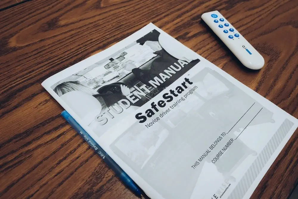 SafeStart-student-manual-DriveWise A student manual titled "SafeStart Novice driver training program" lies on a wooden table next to a remote control and a blue pen. The manual cover features black and white images of people in a car.