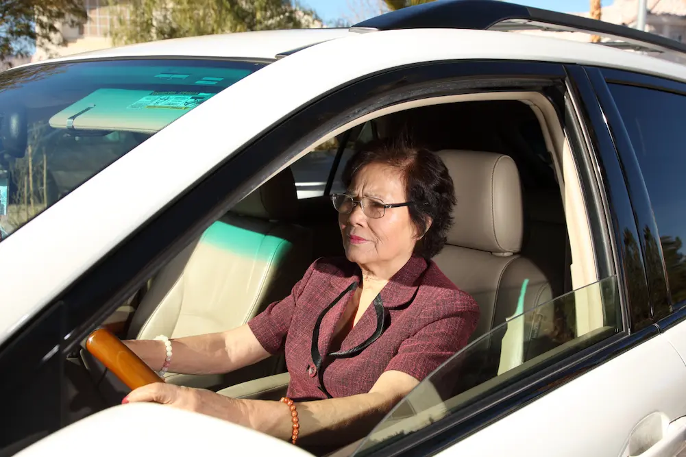 Senior-driver-DriveWise An older woman wearing glasses and a maroon blouse is sitting in the driver’s seat of a white car, holding the steering wheel and looking forward. Sunlight is shining into the vehicle.