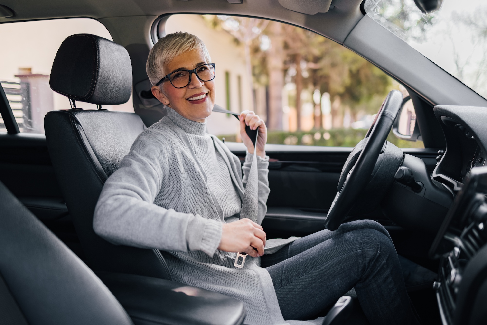 Portrait of happy senior woman fastening seat belt before driving a car. A person sits in a driving simulator with three large screens showing a virtual urban street scene with vehicles, pedestrians, and buildings. The person holds a steering wheel and looks at the central screen.