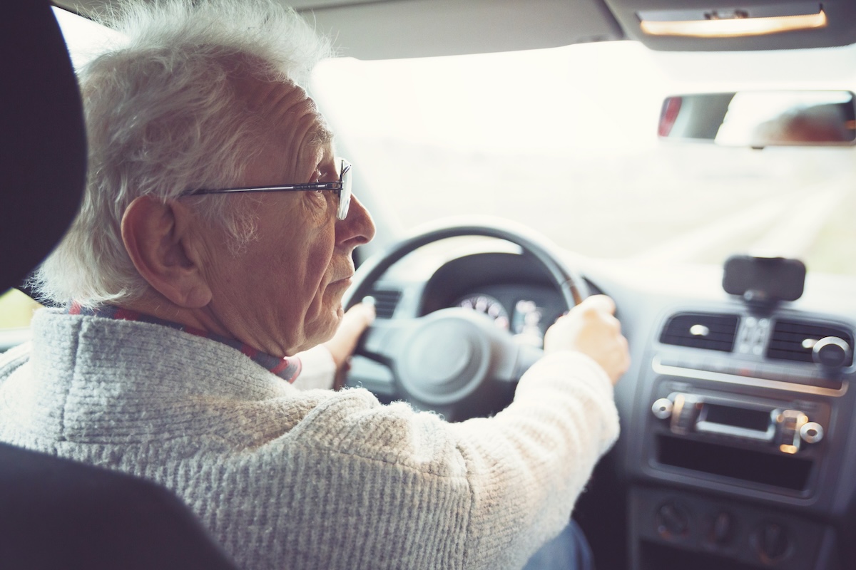Old gentleman drives car An older man with gray hair, wearing glasses and a sweater, sits in the driver's seat of a car, looking over his shoulder while holding the steering wheel with both hands.