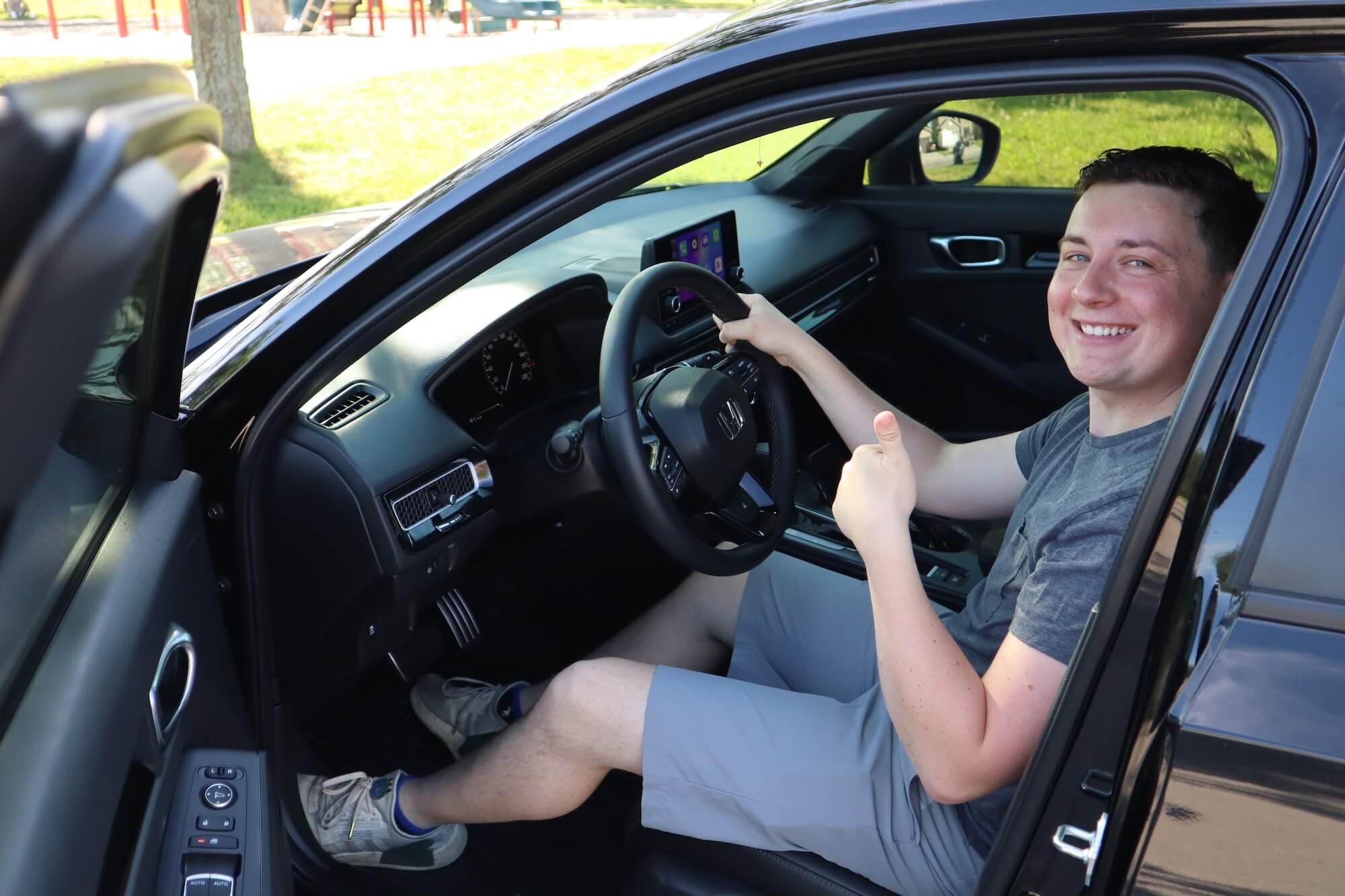 Student-giving-a-thumbs-up-DriveWise-Newmarket A person wearing a gray t-shirt and gray shorts sits in the driver's seat of a car, smiling and giving a thumbs-up. The car door is open, and there is daylight outside.