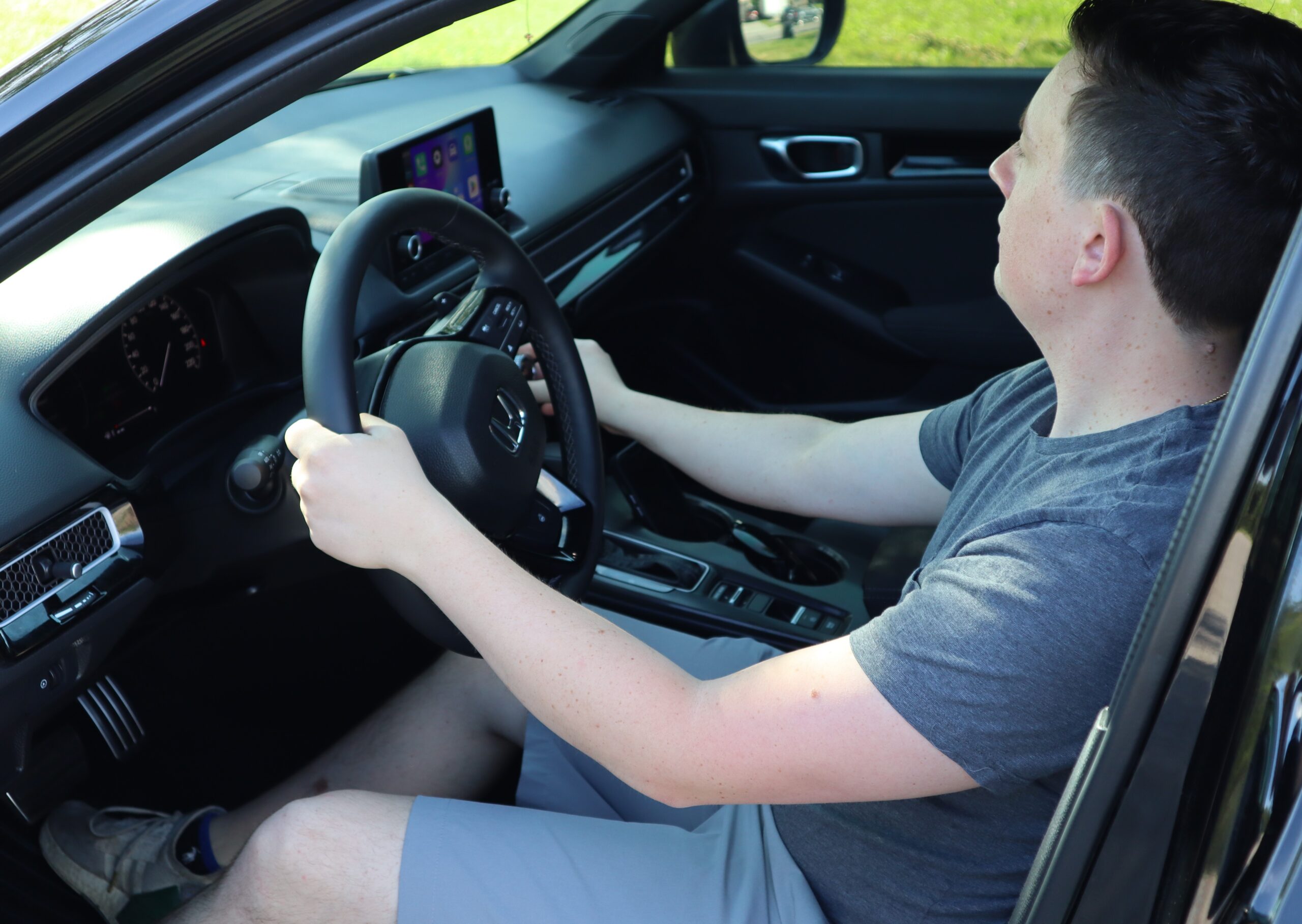 AdjustingAC A person in a gray t-shirt and shorts sits in the driver's seat of a car, holding the steering wheel with both hands and looking forward. The car's dashboard and infotainment system are visible.