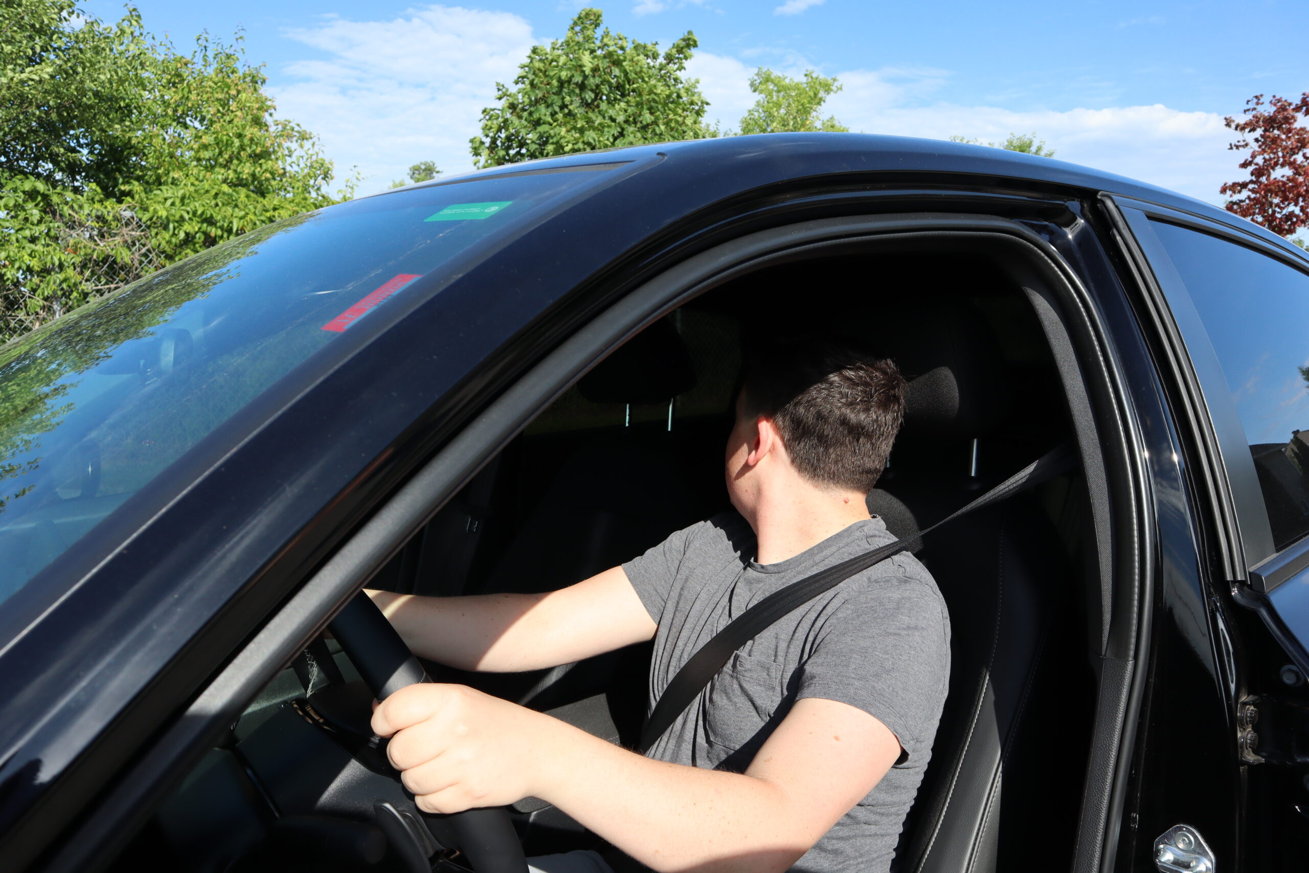 LookingBack A person wearing a seatbelt is sitting in the driver's seat of a car, looking over their right shoulder while holding the steering wheel. The car is parked outdoors with trees and a blue sky visible.